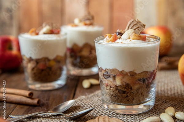 Fototapeta Kuchen im Glas mit Stücken vom Bratapfel, Zimt, Spekulatius, Apfel, Mandeln und Rosinen mit einer Creme aus Quark oder Mascarpone auf einem Tisch aus Holz in einer gemütlichen Küche zu Weihnachten