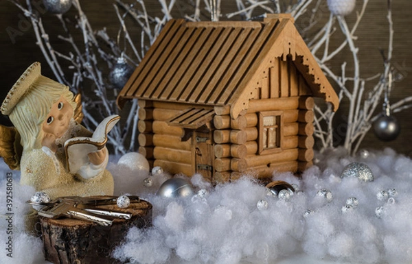 Fototapeta Model of a wooden house with a figurine of an angel, a cut from a tree and house keys in artificial snow on a background of white branches with glass balls. Christmas composition.