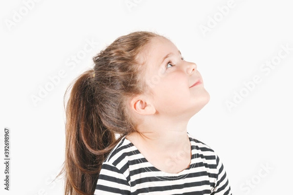 Fototapeta Side profile view of cute smiling girl looking up aside. Girl is waiting for something amazing, looking and listening with attention. Posing little girl wearing striped shirt.