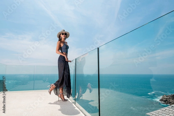 Obraz Fashionable girl with stylish accessories in a hat, a long skirt posing on a transparent balcony overlooking the sea and rainforest