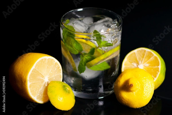 Fototapeta In the center of the screen are a glass of water, ice cubes, a sprig of mint and a slice of lemon. Cut lemons lie to the right and left. Closeup.