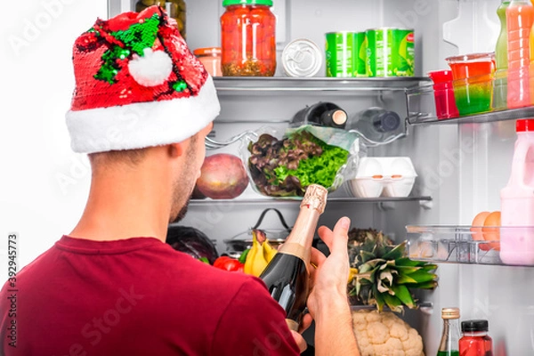 Obraz A man in a Christmas hat holds a bottle of champagne against the background of an open refrigerator with food. Concept for new year or party