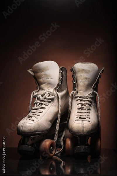 Obraz Old heavily used worn-out pair of white roller skates showing their boot tongues with some dirt and untied shoelace on reflective surface and warm brown background with upper space for titles