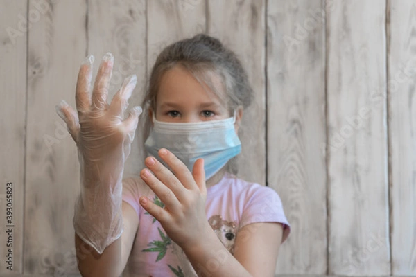 Fototapeta A little girl with a medical mask put on disposable gloves and admires how well she did, spoiling the coronavirus, in a pink t-shirt, can't put on a glove, wooden background photos