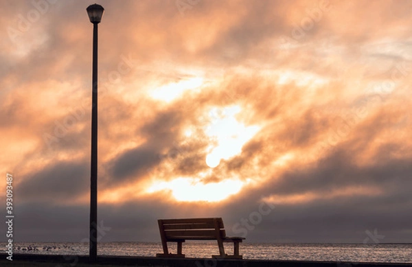 Fototapeta A Wooden bench and street lamp with a background view of a  bright sunset