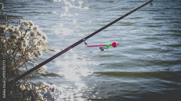 Fototapeta Fishing rod with a bell on the lake in autumn against the background of sun glare on the water.