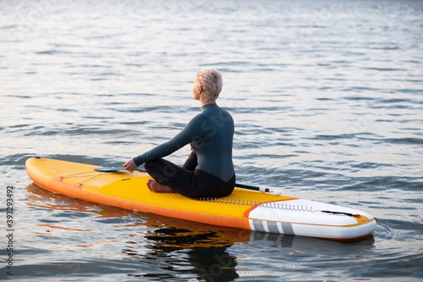 Obraz A young athletic woman in a blue wetsuit swims through the sea on a yellow paddle Board and meditates in the Golden rays of the dawn sun