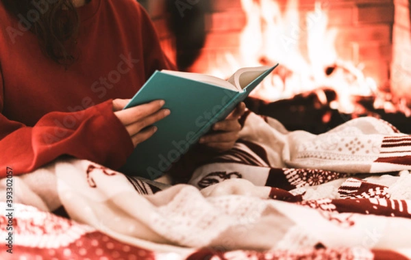 Fototapeta Female resting and warming up in a cold day covering with a blanket and reading a book - festive atmosphere with fire place on background