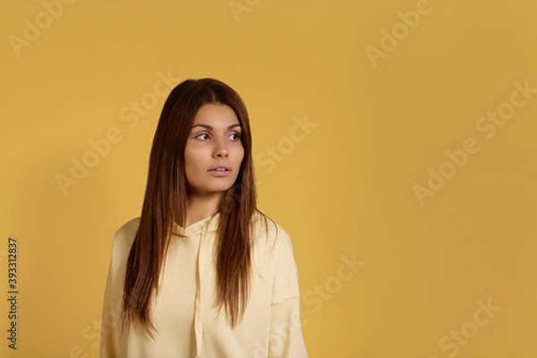 Fototapeta Anything else. Serious and confident caucasian woman in yellow hoodie standing with his back to camera, turned head to the side in anticipation of something. Isolated over a yellow background.
