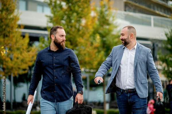 Fototapeta Satisfied business colleagues discussing project on their way to office. Business men walking outside, holding papers, talking, laughing. 