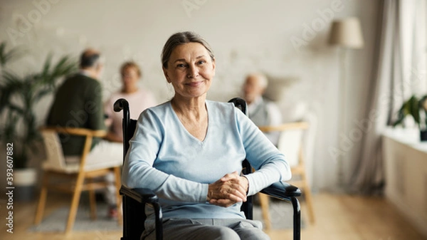 Fototapeta Portrait of disabled senior woman in wheelchair looking at camera and smiling happily in nursing home; other aged patients in background
