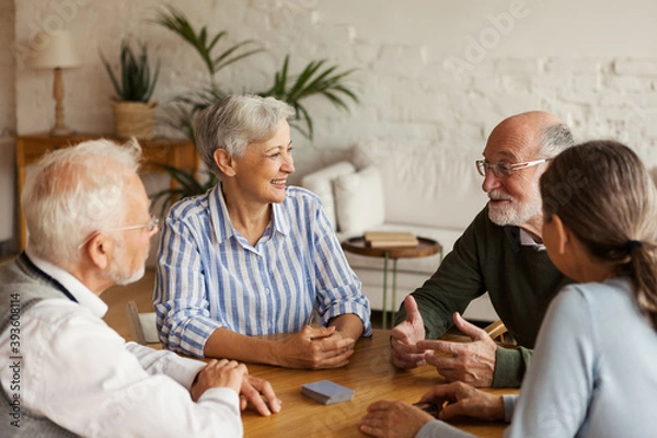 Fototapeta Group of four cheerful senior friends, two men and two women, sitting at table and enjoying talk after playing cards in assisted living home