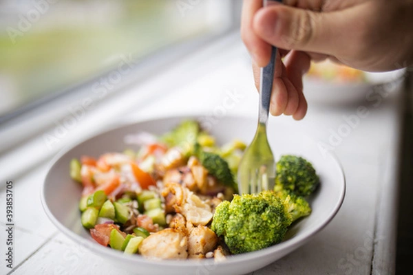 Fototapeta Hand sticking fork on broccoli and chicken salad next to a window
