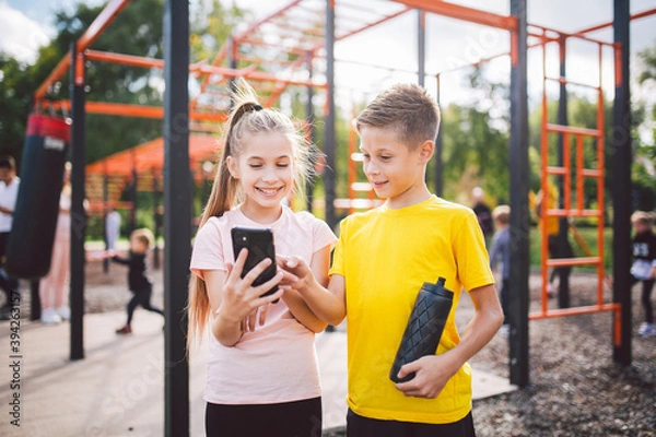 Fototapeta Two children athletes use a smartphone at an outdoor gym. Twins boy and girl are watching the results of the exercise on the phone. Little friends sportsman enjoy the content from the cell phone