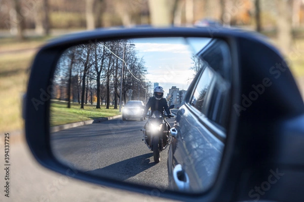 Fototapeta Mutual passing, motorcycle and vehicle with dazzle lighting overtaking the car, view in a side mirror. Improper driving or traffic violation