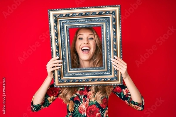 Fototapeta Young caucasian woman with blond hair holding empty frame smiling and laughing hard out loud because funny crazy joke.