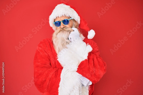 Fototapeta Old senior man wearing santa claus costume and sunglasses serious face thinking about question with hand on chin, thoughtful about confusing idea