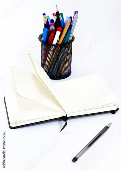 Fototapeta Black pen, open small unfold notebook, note with blank empty light pages near metallic glass stationery with pens and felt pens different colors. White background. Office
