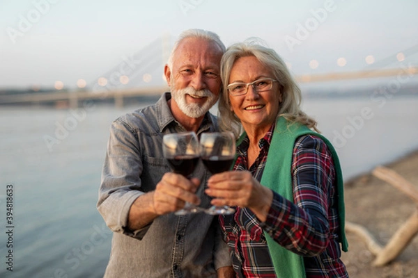 Fototapeta Portrait of happy elderly couple drinking wine on the riverside and toasting. 