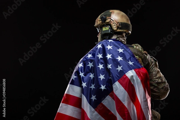 Fototapeta Back view of a military man in a hard hat and full gear, draped over the shoulder of flag of America isolated on black. The concept of patriotism, protection of the Motherland, love for the country
