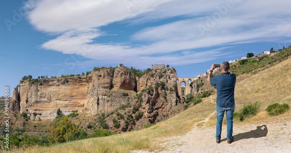 Obraz A male tourist photographs the famous arched bridge of Ronda in Andalusia in summer with a cloudy blue sky. The city is on top of the plateau. At the very bottom is a small waterfall.