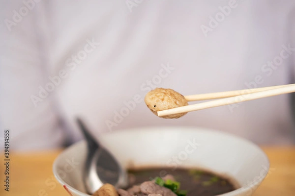 Obraz Unrecognizable Asian woman picking up a meat ball from the noodle bowl by using a wooden chopsticks close up. 