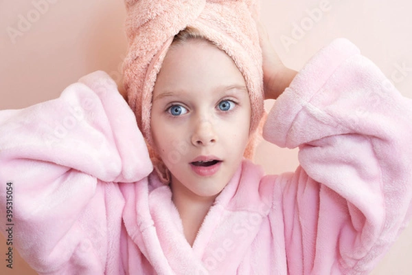Fototapeta a little girl with blue eyes in a pink robe and a pink towel on her head after a bath smiles and looks into the frame. on a peach background