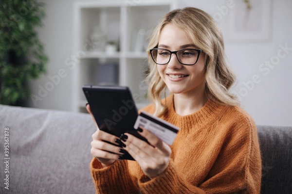 Fototapeta Happy young woman in eyeglasses using digital tablet and credit card for online shopping while staying at home. Concept of modern technology.