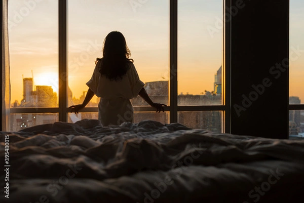 Obraz Beautiful asian woman is waking up in the morning, standing near bed, Sun shines on her from the big window. Happy young girl greets new day with warm sunlight flare and city scenery in the window.