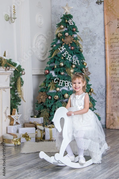 Obraz little girl riding a wooden horse near the fireplace and New Year tree