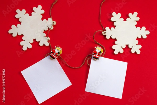Obraz New year's holiday background. White sheets for notes on wooden clothespins with figures of Santa Claus and white empty wooden snowflakes on a red background. Flat lay. Greeting card for Christmas.