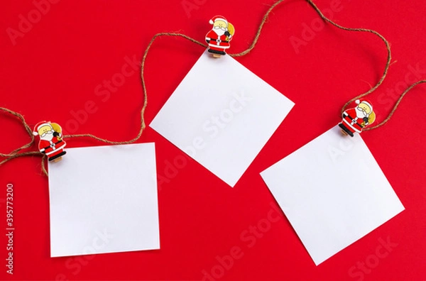 Obraz New year's holiday background. White sheets for notes on wooden clothespins with figures of Santa Claus on a red background. The view from the top. Greeting card for Christmas.
