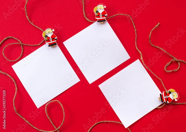 Obraz New year's holiday background. White sheets for notes on wooden clothespins with figures of Santa Claus on a red background. Concept of an eco-friendly holiday. Flat lay. Greeting card for Christmas.