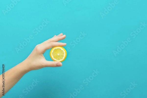 Fototapeta Female hand holding lemon fruit slice on blue background with copy space