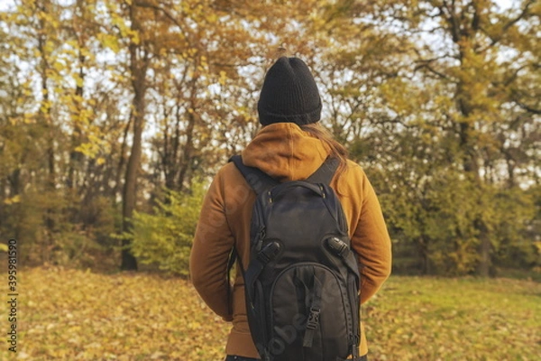 Fototapeta Woman in a yellow jacket with her hends in her pockets and a backpack on her back walking trough the woods in the fall