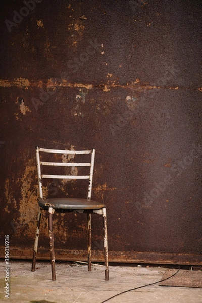 Fototapeta A rusty chair with peeling paint and a shabby leatherette seat against a rusty wall on the concrete floor.