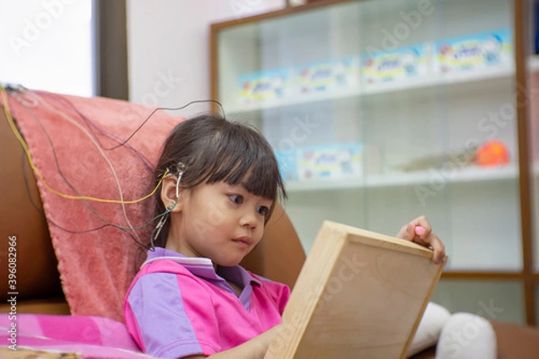 Fototapeta A little girl is wearing a hearing aid and is doing a neurofeedback exam. Making EEG electrodes.