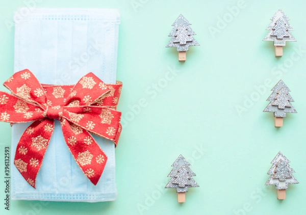 Obraz medical protective face masks as a gift with a red ribbon on a green background. Christmas and New Year 2021 decor painted firs and snowflakes. Holidays self-isolation and coronavirus pandemic concept