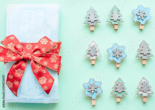 Obraz medical protective face masks as a gift with a red ribbon on a green background. Christmas and New Year 2021 decor painted firs and snowflakes. Holidays self-isolation and coronavirus pandemic concept