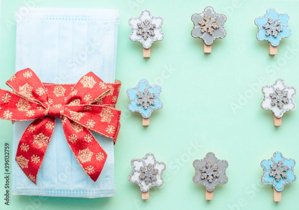 Obraz medical protective face masks as a gift with a red ribbon on a green background. Christmas and New Year 2021 decor painted firs and snowflakes. Holidays self-isolation and coronavirus pandemic concept