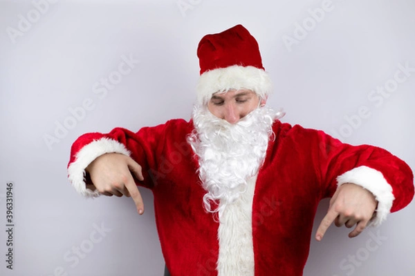 Fototapeta Man dressed as Santa Claus standing over isolated white background surprised, looking down and pointing down with fingers and raised arms