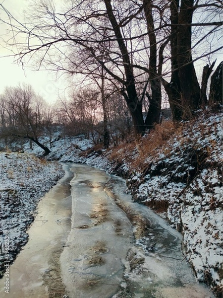 Fototapeta frozen small river with snow capped banks against the backdrop of trees with fallen leaves