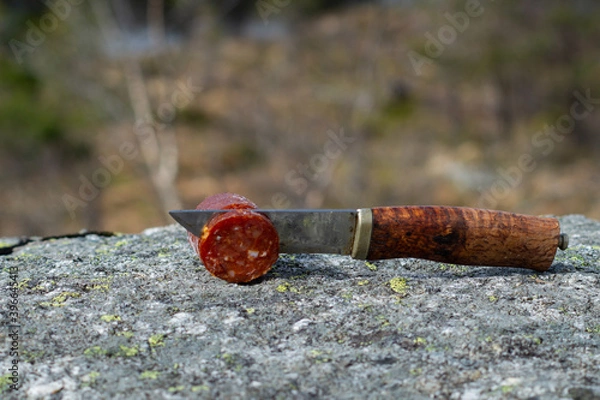 Fototapeta Knife cutting sausage in the mountain with blurry mountain landscape in background.  Outdoor food.