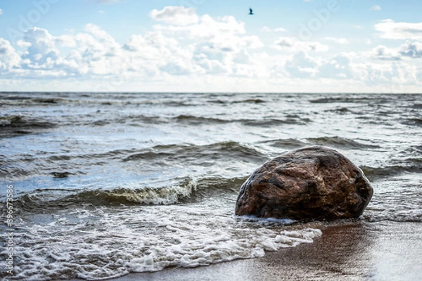 Fototapeta The wave beats against a large stone on the seashore, a clear day, light clouds