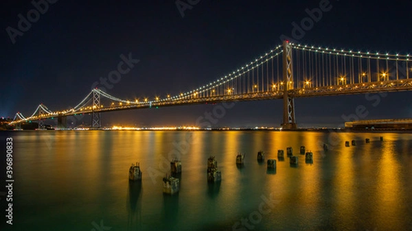 Fototapeta Night image of the Oakland Bridge as it leaves San Francisco and heads northwest into Oakland with lighted reflections of sunken pier ballasts