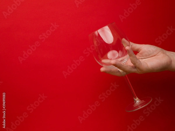 Fototapeta Hand holding an empty wine glass on red background. Side view with space for copying. Concept of holiday backgrounds.