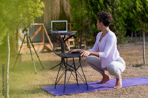 Obraz Yoga trainer holding online class during pandemic Coronavirus. Fitness coach teaching yoga online to group of people near the sea side in hotel garden. working outside, untethered employers, gig econo