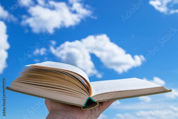 Obraz An open book in a hand raised to the sky. Blue sky with clouds create a background for the book.