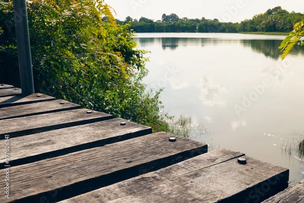 Fototapeta Close up view of rustic wooden bridge above the pond against reflection of sky and trees on calm water background.