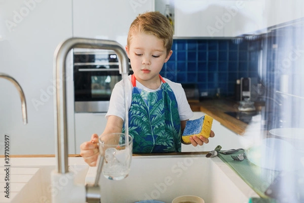 Fototapeta a boy in an apron washes dishes in the kitchen at home.
